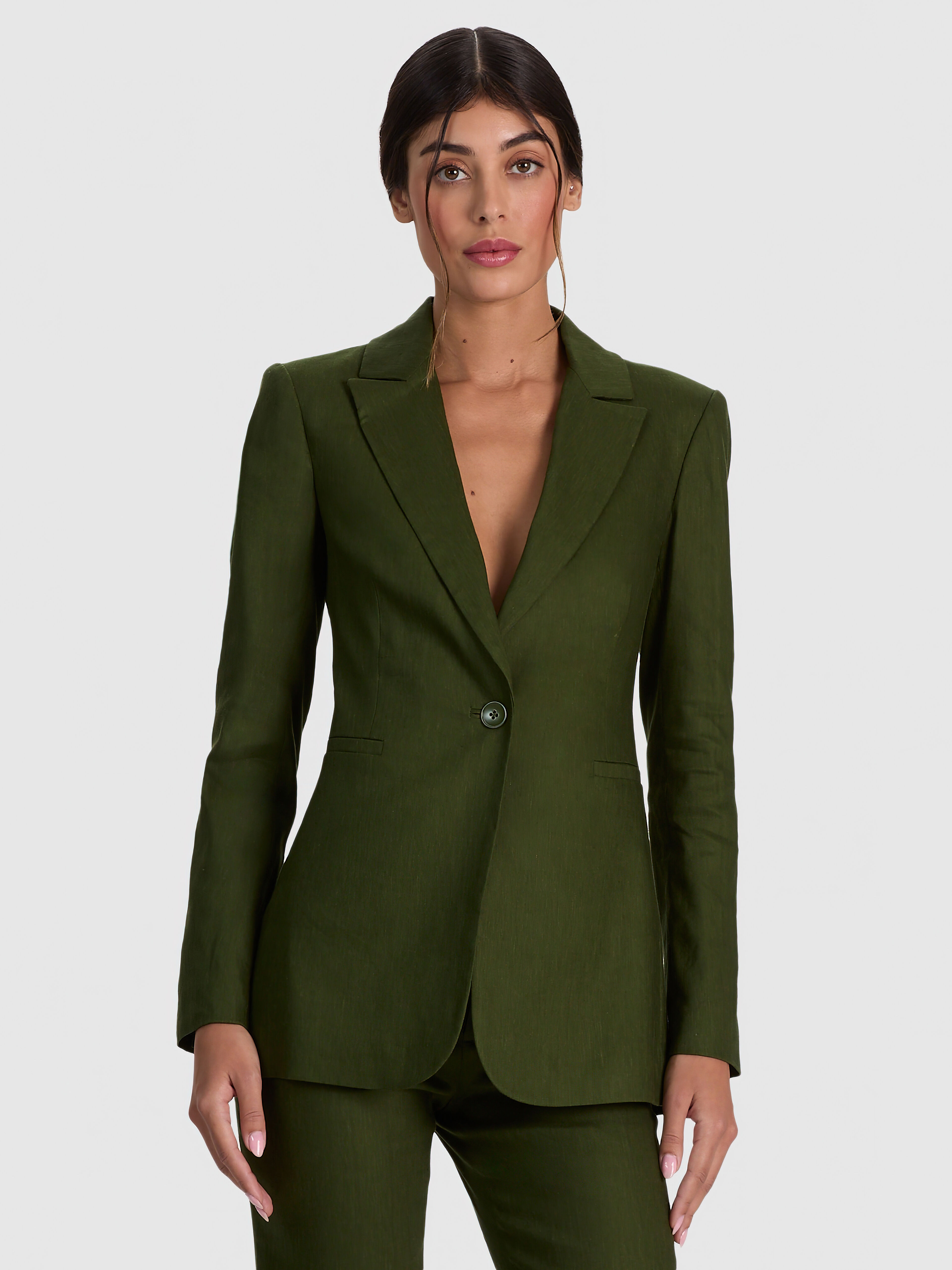 A campaign-style editorial image of a woman wearing the ALICE + OLIVIA Breann Linen Blazer in dark green, styled as a sharp tailored suit with matching trousers against a clean backdrop.