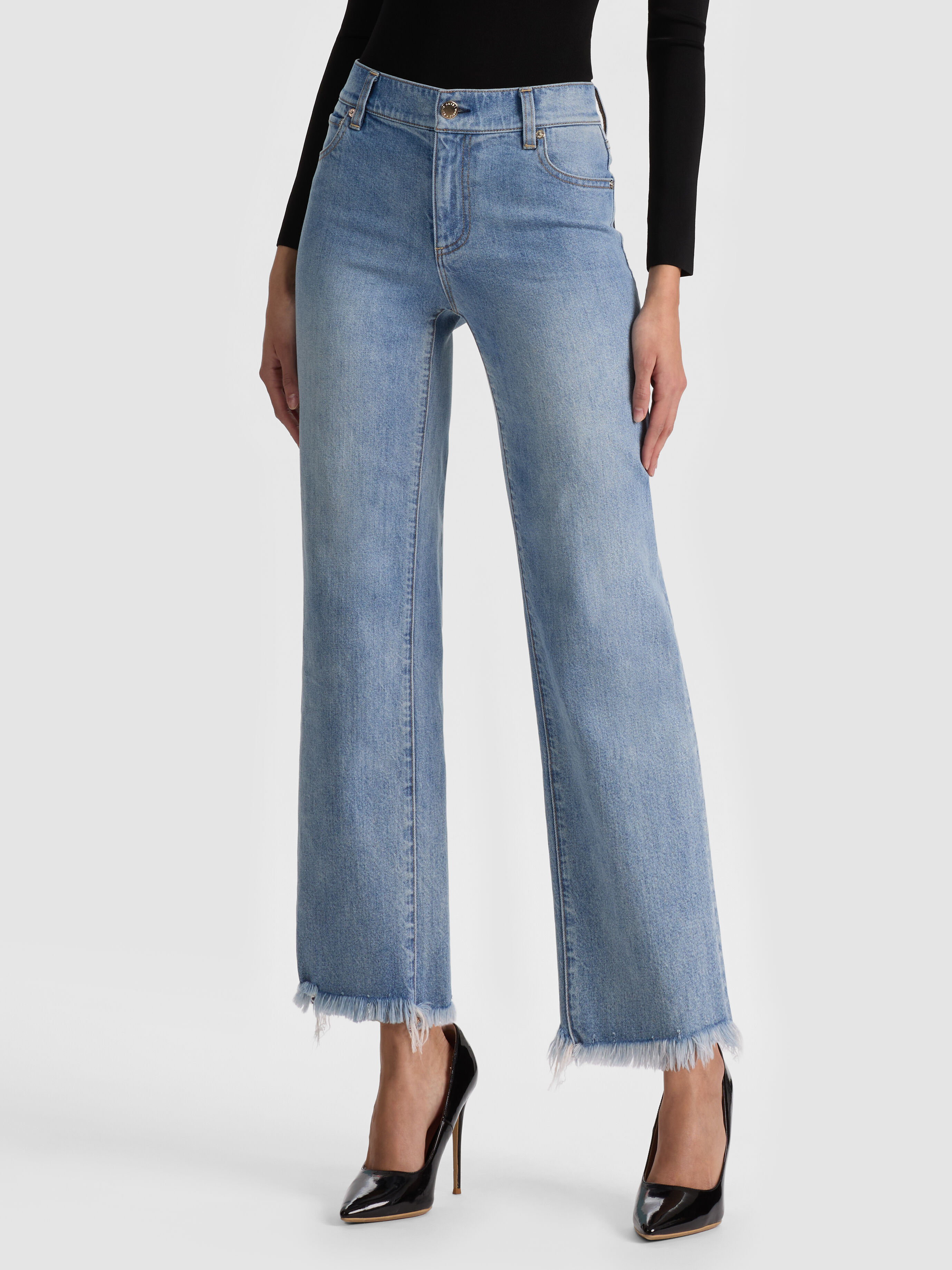 A full-length lifestyle shot of a woman wearing the ALICE + OLIVIA Fierce Jean, a classic slim straight-leg silhouette with a raw hem, styled with heeled boots, showcasing the timeless straight-leg staple denim trend for 2026.