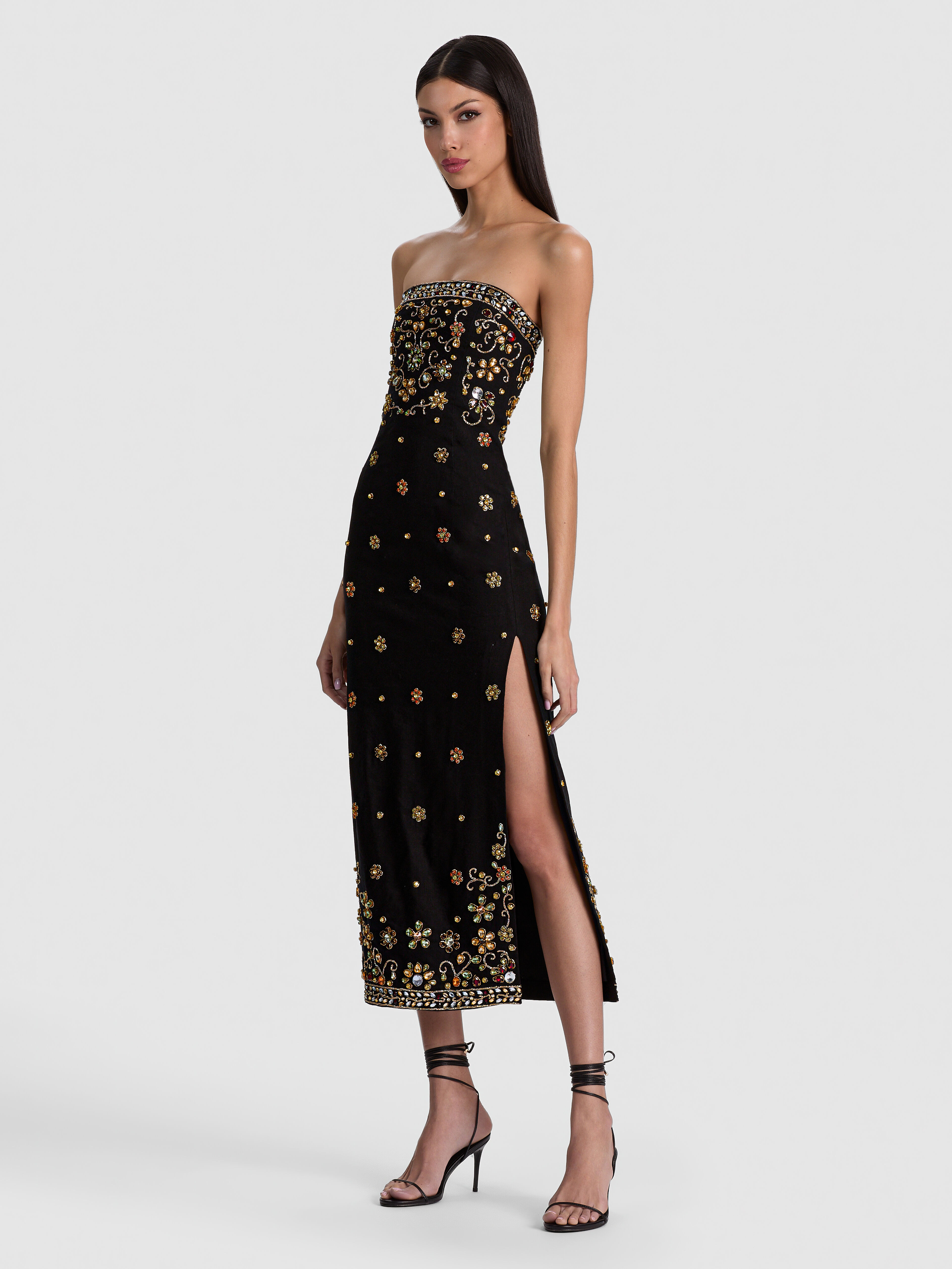 A model wearing the ALICE + OLIVIA Nina Embellished Midi Dress in a strapless black silhouette with intricate multicolored crystal embroidery, styled with statement earrings and strappy heels for a formal evening event.